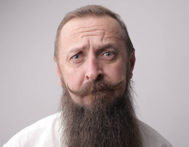 istock Man with a long beard and a mustache frowning while standing in front of a grey wall 1439794940