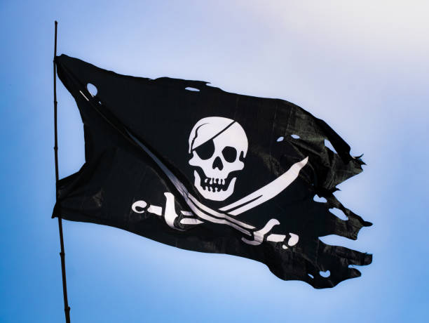 istock Completely torn and bruised black colored pirate flag (Jolly Roger) fastened to a flagpole flutters in the wind with a skull with patch and two swords on the bottom in white drawn on the fabric 1439798926