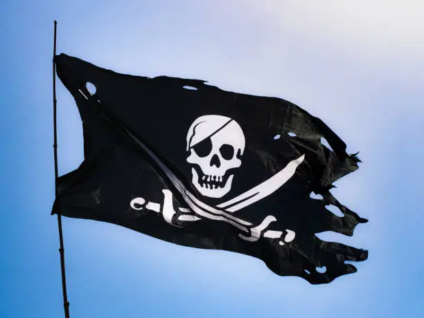 Photo of Completely torn and bruised black colored pirate flag (Jolly Roger) fastened to a flagpole flutters in the wind with a skull with patch and two swords on the bottom in white drawn on the fabric