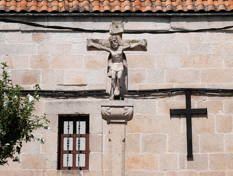 Stone monument of a crucified Christ in the center of a square of a Galician city illuminated by the sun and a background facade of a church with a black metal cross and a window with bars