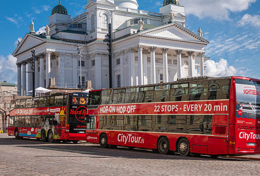 Helsinki, Finland - July 19, 2022: Closeup of red Hop-on Hop-off public buses on Senate Square with Cathedral in back under blue cloudscape.