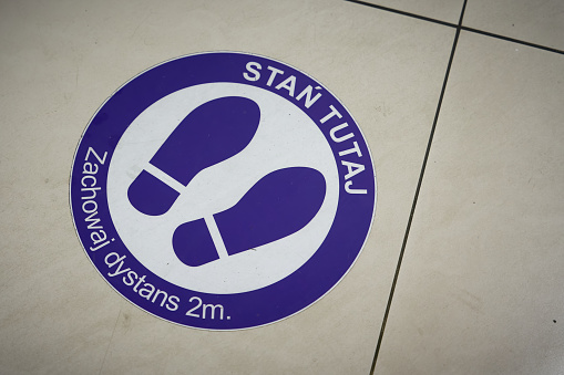 Sticker on the floor of a shopping center in Poland with the words: Stan Tutaj Zachowaj dystans 2m. Translation: Keep a distance of 2m