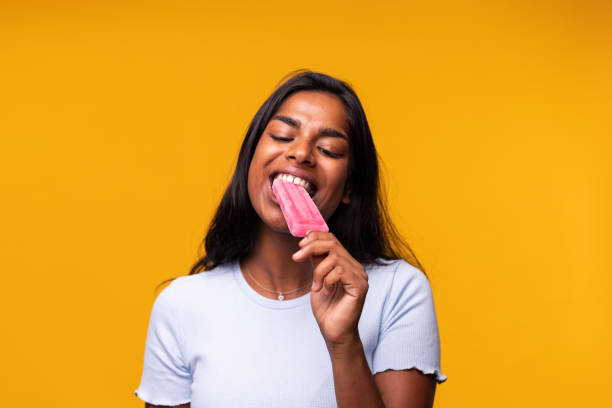 Young Indian woman eating pink popsicle on yellow background. Asian woman eating ice cream. Young Indian woman eating pink popsicle on yellow background. Asian woman eating ice cream. Studio shot. popsicle stick stock pictures, royalty-free photos & images