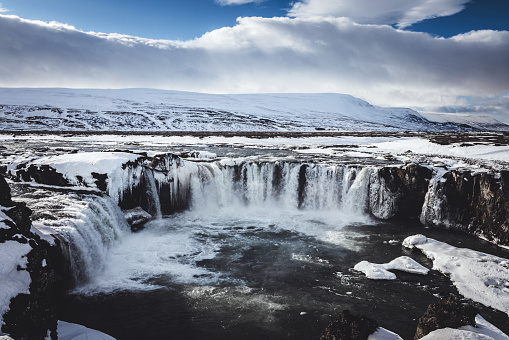 Iceland Godafoss Waterfall in Winter. View towards the famous Godafoss Falls -  icelandic “Waterfall of the Gods” under sunny winter skyscape, snow covered Mountains in the background. The ice cold water of the river Skjálfandafljót falls from a height of 12m over a width of 30m. Akureyri, Fossholl, Northern Icelandic Highlands, Iceland, Nordic Countries, Europe