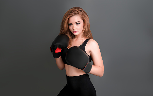 A young girl in boxing seals, a black top with bare shoulders, leggings and brown hair, stands in a protective pose on a gray background with copy space.
