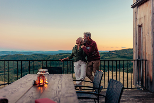 An affectionate senior Caucasian couple is at their mountain getaway spot enjoying the beautiful sunset in the nature on the deck.