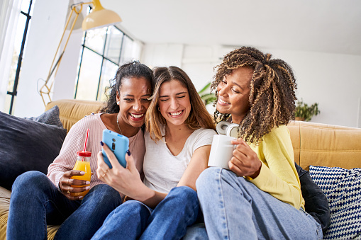 Indoor happy woman lifestyle portrait of three funny young female friends have fun and pretending smiling faces. Home party mood girls using mobile phone taking selfie photo. High quality photo