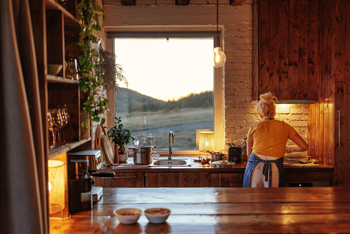 A woman is in her modern but rustic kitchen preparing a meal with a beautiful view of the mountain through the window.