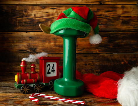 Heavy green dumbbell with elf hat. Healthy fitness lifestyle holiday season concept with toy train advent calendar, candy cane and Santa Claus hat. Exercise equipment as a Christmas gift idea. Gym workout sport training composition on wooden background.