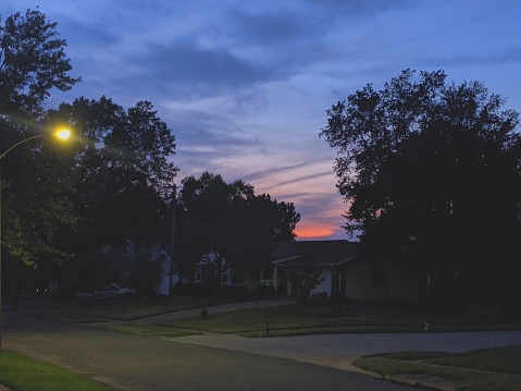 Suburban neighborhood at sunset with a wide empty street and a line of houses in shadow below dark trees and a blue and orange sky.