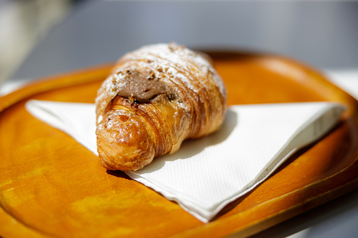 Croissant with chocolate topping iced with powder sugar, served on napkin on wooden board