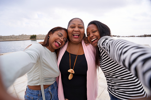 Cheerful portrait of three African American women taking selfie looking at camera with big smile, Family of three enjoying by the sea outdoors. One middle age woman and two young adult girls.