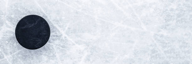 istock Black old rubber puck on ice background. Closeup. Wide banner. Top down view. Empty place for text. 1439823901