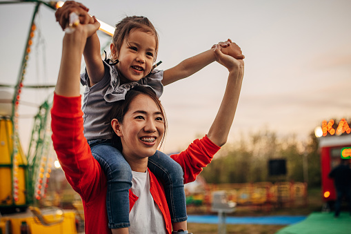 Two people, mother carrying her cute little daughter on shoulders, they are having fun together in amusement park.