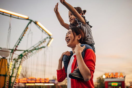 Two people, mother carrying her cute little daughter on shoulders, they are having fun together in amusement park.