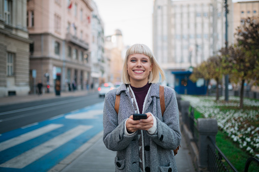 Portrait of a happy young adult woman with blond hair standing on a bus stop in the city, using a smart phone to call for Uber while looking at the camera