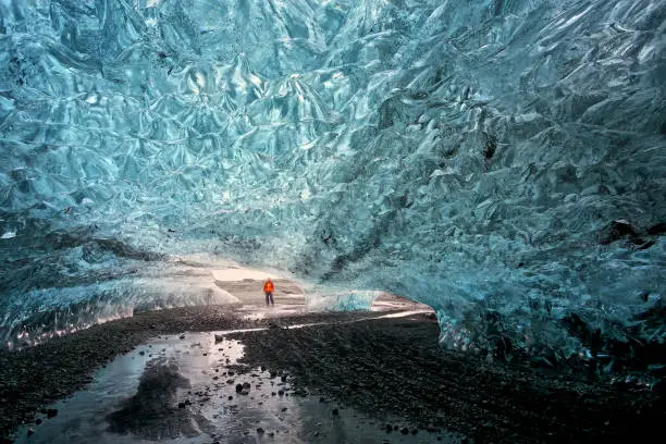 Man with an orange jacket standing in front of the entrance of a glacier in Iceland. Shot from inside the cave, the man is looking at the camera. Cave under a glacier with a blue translucent ceiling