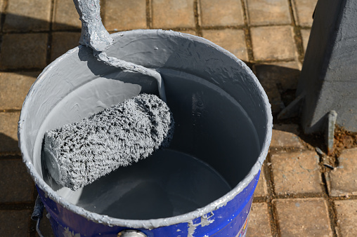 A paint roller in a metal bucket with gray paint on the street. The process of updating the streets of the city