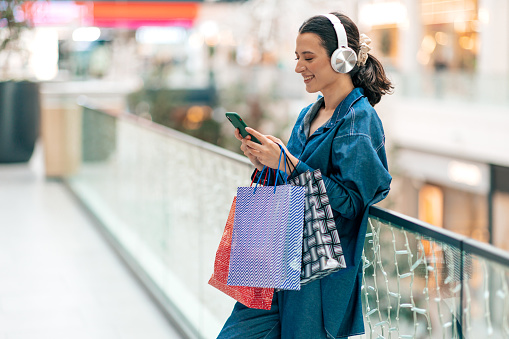 Beautiful young Caucasian woman with a bright smile on her face standing with headphones in shopping mall and using smart phone
