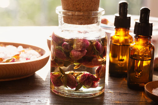 aromatherapy oil in a glass bowl bottle with dried roses and almond oil, surrounded by wooden bowls with sea salt and brown pipette bottles