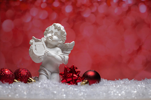 Ceramic angel statue and christmas tree toys of red color standing on snow on red bokeh background.Copy space.Xmas greeting card concept.Christmas background.