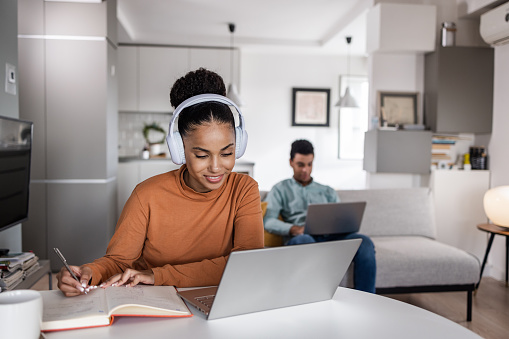 Young multiracial couple working at home in the same room. They are concentrating on the work. Woman is sitting at the table and writing notes while listening something on the laptop. Man is sitting on the couch.