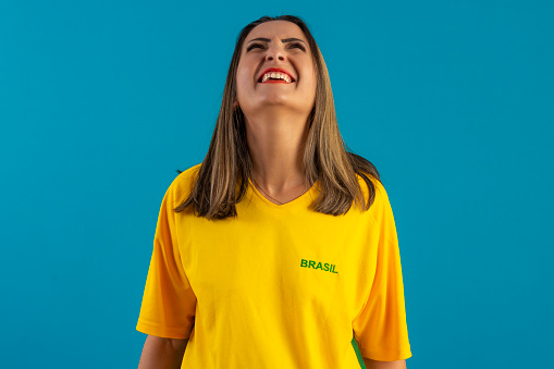 Brazilian female supporter, wearing shirt and Brazilian flag. Brazilian soccer fan in studio photo with blue background and Brazil colors.