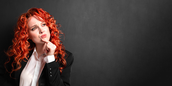 Portrait of a red-haired woman entrepreneur in a black jacket and white shirt thinking on a dark background. Concept idea and planning.