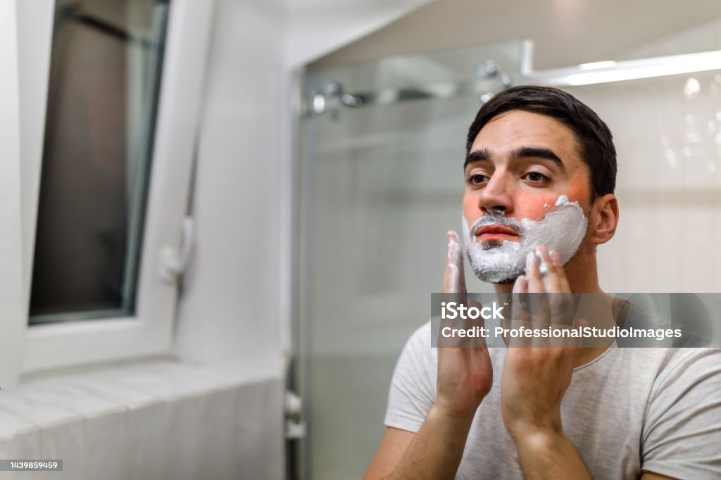 A man is preparing for shaving in the bathroom by applying shaving foam. A young man is applying shaving cream in front of a mirror in a bathroom and preparing to shave his beard. Morning Stock Photo