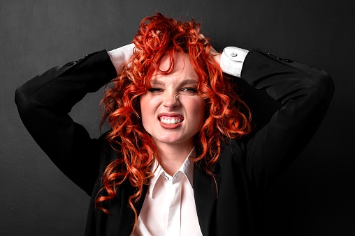 A red-haired woman in a black jacket and a white shirt clutches her head in hysterics. A portrait of a woman with expression and emotions. The concept of a panic attack, mental health.