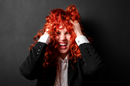 A red-haired woman in a black jacket and a white shirt clutches her head in hysterics. A portrait of a woman with expression and emotions. The concept of a panic attack, mental health.