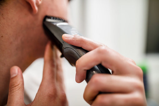 A man is standing in front of the mirror in the bathroom and shaving his beard with an electric razor.