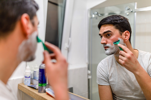 A man is standing in front of the mirror in the bathroom and shaving his beard with a razor.