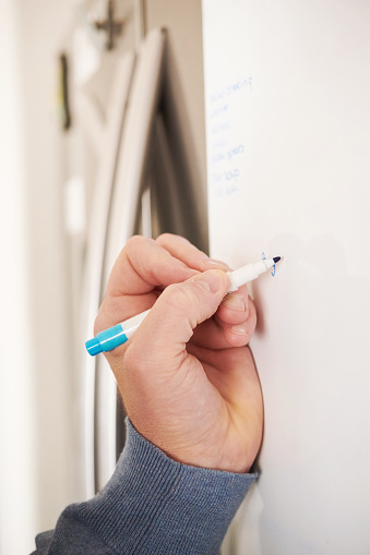 Unrecognizable middle aged man writing a shopping list on a whiteboard on a kitchen refrigerator
