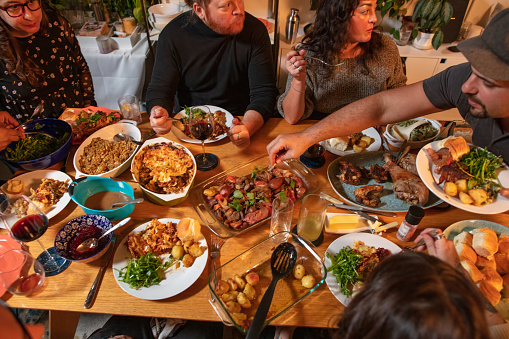 High angle view of a group of people gathered around a table in a loft apartment for a friendsgiving dinner, reaching across one another to serve themselves.