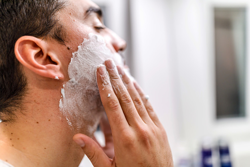 A young man is applying shaving cream in front of a mirror in a bathroom and preparing to shave his beard.