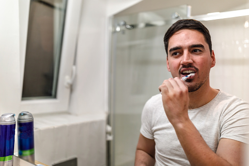 A man is standing in front of the mirror in the bathroom and brushing his teeth using a toothbrush.