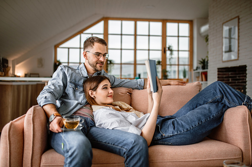 Young couple sitting in their living room in sofa, reading book and relaxing together.
