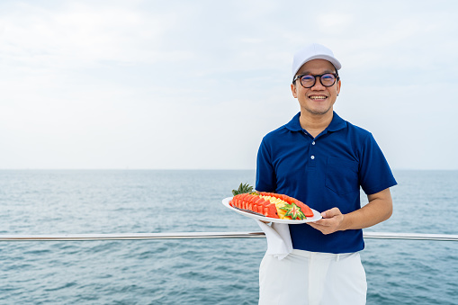 Portrait of Happy Asian man waiter holding fresh fruit on serving dish for serving to passenger tourist travel on luxury catamaran boat yacht on summer vacation. Cruise ship service occupation concept