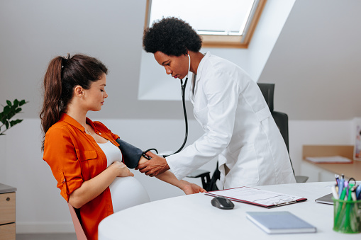 A mid adult African American doctor is measuring the blood pressure of a young Caucasian pregnant woman in her office.
