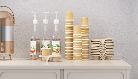 Close up stacks of disposable paper cups and snack bowls with stainless steel hot water container with variety of syrup pump bottles on pantry counter. Space, products display backdrop.