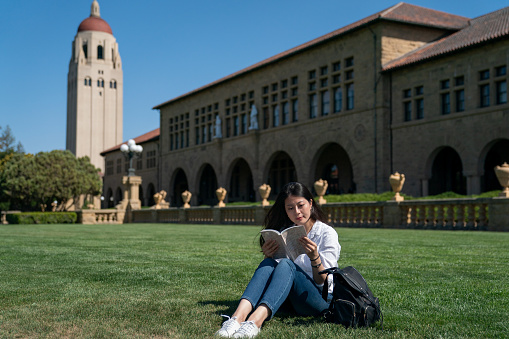leisure asian female exchange student enjoying reading while sitting on green lawn in front of hoover tower during break time on a sunny day at school university in California usa