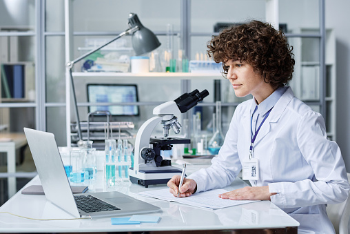 Young female scientist in labcoat making notes while sitting by workplace in front of laptop in laboratory and watching online video