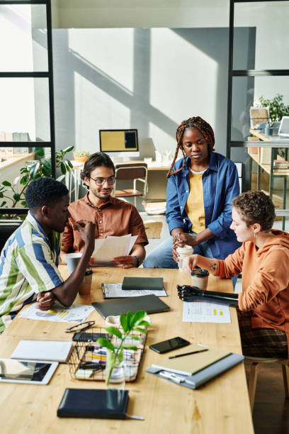 Group of young employees gathered by table in office Group of young employees gathered by table in office to have coffee and discuss some working points concerning new business project workplace diversity vertical stock pictures, royalty-free photos & images
