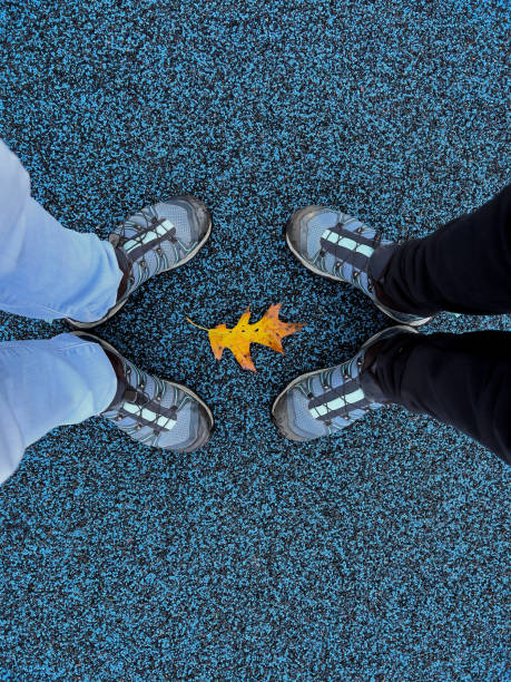 Mature Asian mother and Eurasian adult daughter wearing the same shoe style and standing on blue recycled rubber at and outdoor playground with an orange autumn leaf framed between their feet.  North Vancouver, British Columbia, Canada.