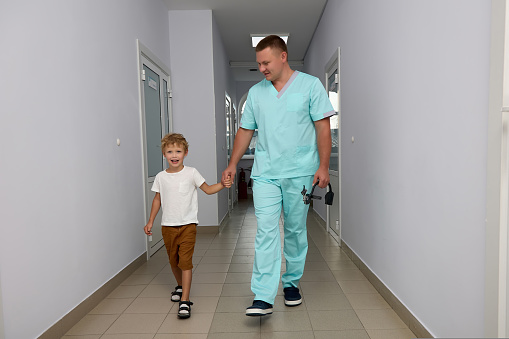 Pediatrician with a medical instrument in his hand leads a little boy by the hand to the examination room. Smiling boy goes with a doctor by the hand for a preventive examination