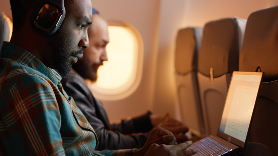 African american passenger in airplane seat flying on work trip, using laptop on flight. Travelling with commercial airline during sunset, wearing headphones on aviation jet.