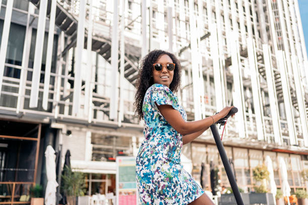 Young black girl wearing colorful dress riding her electric scooter in the city during sunny day.
