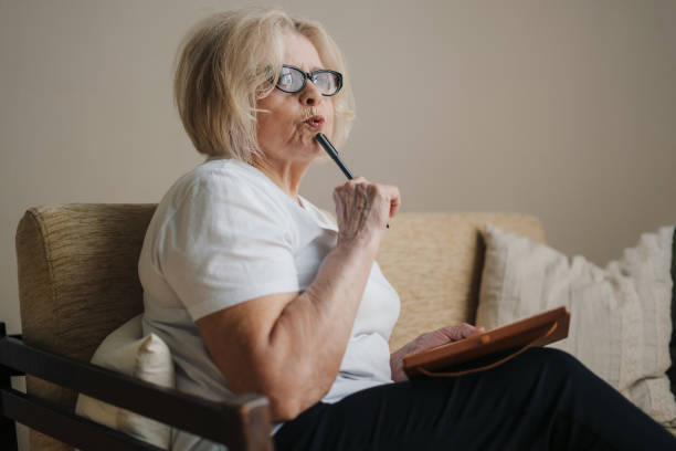 istock Pensive old woman sitting on sofa, looking out the window, thinking about retired life, making notes. Portrait for lifestyle design. Isolating during coronavirus covid 19 quarantine lockdown. Senior woman with pen making notes in notebook. 1439923634