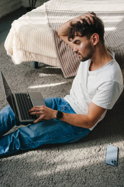 Man with under eye patches working on laptop in bedroom floor at home. Daily skincare beauty routine. Cosmetic concept. Skincare concept. Facial skincare. hombre con parches debajo de los ojos trabajando en una computadora portátil en el piso del dormitorio en casa. rutina diaria de belleza para el cuidado de la piel. concepto cosmético. concepto de cuidado de la piel. cuidado de la piel facial.